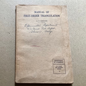 May include: A tan-colored book titled "MANUAL OF FIRST-ORDER TRIANGULATION" with the author's name, C.V. HODGSON, and the department name, "Experimental Department, U.S. Naval Net Depot, Liburon, Calif.", written in cursive. The book also has a stamp that reads "DEPARTMENT OF COMMERCE U.S. COAST AND GEODETIC SURVEY."