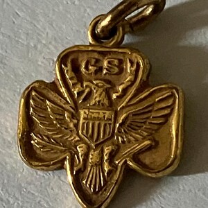 May include: A gold-colored pendant in the shape of a trefoil, featuring an eagle with outstretched wings and a shield. The letters "GS" are visible at the top. The pendant has a small loop for attaching to a chain or bracelet.