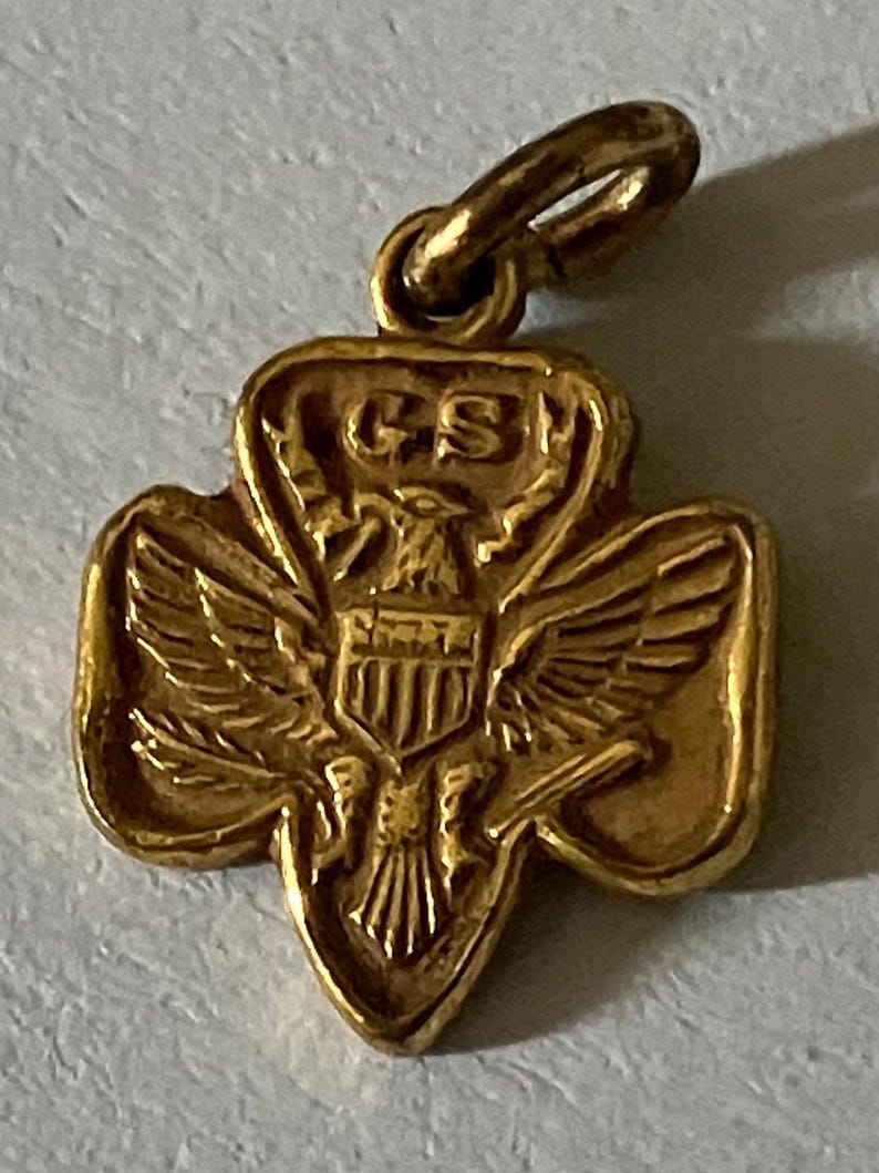May include: A gold-colored pendant in the shape of a trefoil, featuring an eagle with outstretched wings and a shield. The letters "GS" are visible at the top. The pendant has a small loop for attaching to a chain or bracelet.