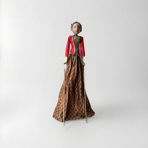 May include: A traditional Indonesian Wayang Golek puppet. The puppet is dressed in a red jacket with green trim, a brown patterned skirt, and a carved wooden head. The puppet is supported by wooden sticks.