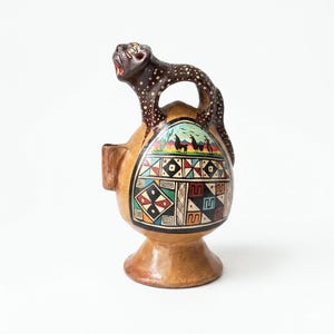 May include: A brown ceramic vessel with a decorative design. The vessel features a handle and a spout. A dark brown spotted animal figure is perched on top. The body of the vessel is adorned with colorful geometric patterns and a landscape scene.