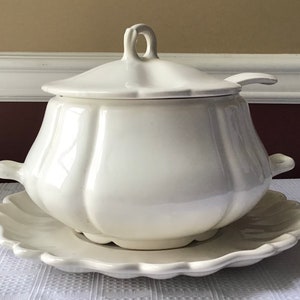 May include: White ceramic soup tureen with a lid and a matching serving plate. The tureen has a fluted design and a handle on each side. The lid has a small handle on top.