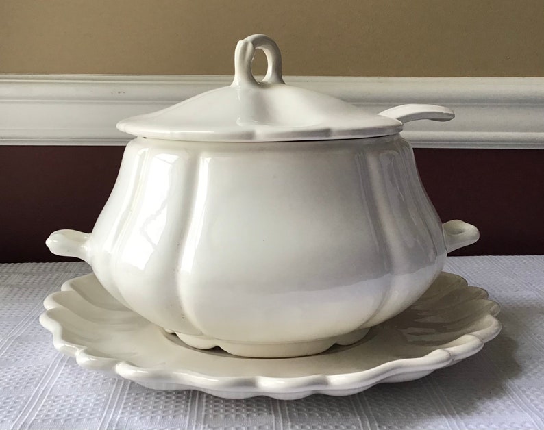 May include: White ceramic soup tureen with a lid and a matching serving plate. The tureen has a fluted design and a handle on each side. The lid has a small handle on top.