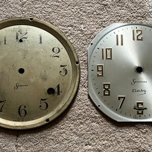 May include: Two vintage clock faces. One is a gold-toned metal with black numerals and the word "Sessions". The other is silver-toned with gold numerals and the words "Sessions Electric". Both have a central hole.