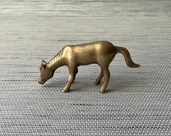 May include: A small, gold-colored metal figurine of a horse with its head down as if it is grazing. The horse is standing on a gray textured surface.