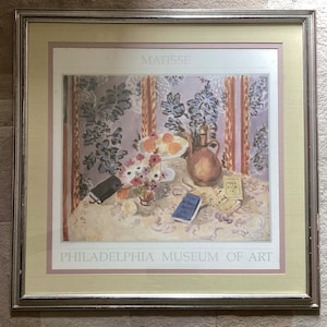May include: A framed print of a still life painting by Henri Matisse. The painting depicts a table setting with a pitcher, fruit, flowers, and books. The title "MATISSE" is printed at the top of the print, and "PHILADELPHIA MUSEUM OF ART" is printed at the bottom.