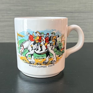 May include: A white ceramic mug featuring a colorful illustration of several people riding a single horse. The scene includes a sign labeled WIDECOMBE and the text WIDECOMBE FAIR printed at the bottom. The mug has a curved handle and a smooth finish.