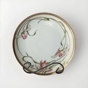 May include: A white porcelain bowl with a gold rim and a floral design. The bowl features pink and green flowers with a stylized art nouveau design.
