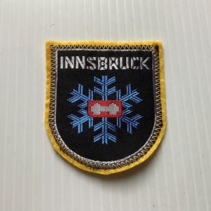 May include: Embroidered patch with the word "INNSBRUCK" in white letters above a blue snowflake design on a black background. The snowflake has a red center. The patch has a yellow border.