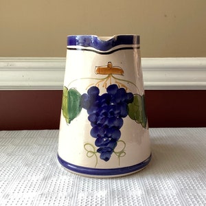 May include: White ceramic pitcher with a blue rim and a painted design of a bunch of purple grapes with green leaves.