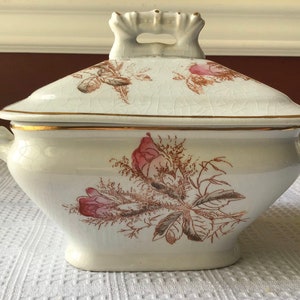 May include: Cream-colored, lidded serving dish with a floral design in pink and brown. The dish has a gold rim and handles on the sides. The lid also features a floral pattern. The dish is likely vintage or antique.