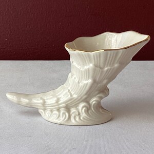 May include: A white ceramic cornucopia vase with a gold rim. The vase is shaped like a horn with a wavy design and a curled tail.