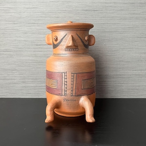May include: A terracotta pottery vase with a human-like face and legs. The vase is decorated with geometric patterns in brown and red. The vase is signed "Panama" on the bottom.