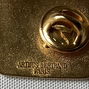 Op de afbeelding: Gouden metalen pin met een getextureerd oppervlak. De achterkant van de pin heeft een veiligheidssluiting en de tekst "ARTUS BERTRAND PARIS" is erop gegraveerd.