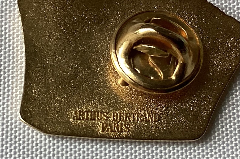 Op de afbeelding: Gouden metalen pin met een getextureerd oppervlak. De achterkant van de pin heeft een veiligheidssluiting en de tekst "ARTUS BERTRAND PARIS" is erop gegraveerd.