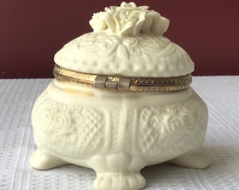 May include: White porcelain trinket box with a floral design and gold trim. The box has a hinged lid and sits on three feet.