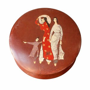 May include: Round, reddish-brown decorative box with a painted illustration of three figures. One figure wears a red dress with gold accents, another a white dress, and the third a purple outfit. The box has a crack on the side.