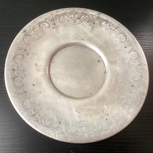 May include: A silver-colored plate with an ornate, engraved border. The plate has a central recessed area and a decorative pattern around the edge. The design features scrolling lines and floral motifs, creating a vintage aesthetic.