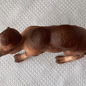 May include: A brown ceramic bear figurine in a crawling position. The bear has a textured fur appearance with varying shades of brown. The figurine is resting on a white, patterned surface.