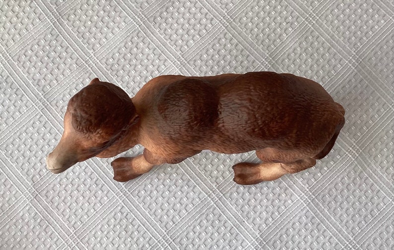 May include: A brown ceramic bear figurine in a crawling position. The bear has a textured fur appearance with varying shades of brown. The figurine is resting on a white, patterned surface.