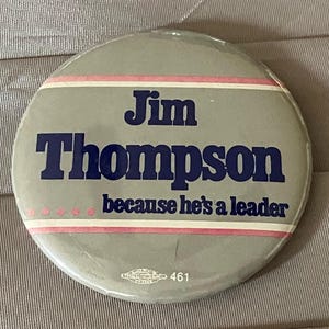 May include: A round, gray button with the name "Jim Thompson" in navy blue, and the text "because he's a leader" below. The button has pink and white stripes and a small logo with the number 461.