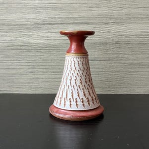 May include: A ceramic candle holder with a red-brown top and base, and a cream-colored body with a textured, linear pattern. The candle holder is on a dark surface.