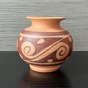 May include: A small, round terracotta vase with a wide mouth. The vase features a band of dark brown, swirling patterns around its midsection. The base and rim are a lighter shade of brown, creating a contrast with the darker design.