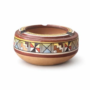 May include: A round, ceramic ashtray with a brown interior and exterior. The ashtray features a geometric pattern in red, yellow, black, and white. The rim has a small notch for resting cigarettes. The ashtray is a decorative item.