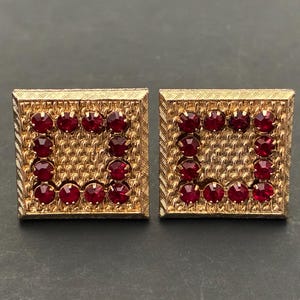 May include: A pair of square gold-tone cufflinks featuring a textured metal border and a central square frame inlaid with twelve small, round, deep red faceted stones arranged in a border pattern against a textured gold-tone background.