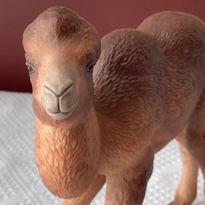 May include: A ceramic camel figurine in shades of brown and tan. The camel has detailed facial features, including dark eyes and a smiling expression. The figurine is standing, with its body and hump visible.