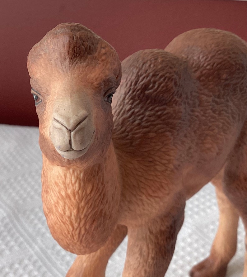 May include: A ceramic camel figurine in shades of brown and tan. The camel has detailed facial features, including dark eyes and a smiling expression. The figurine is standing, with its body and hump visible.