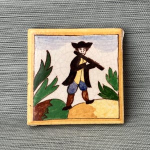 May include: A hand-painted ceramic tile featuring a man in a black coat and hat playing a flute. The tile has a yellow border and a white background with green foliage.