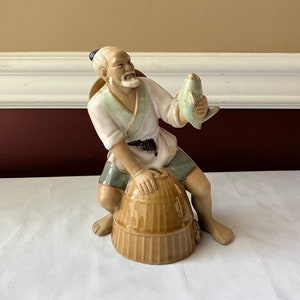 May include: A ceramic figurine of a fisherman holding a fish. The fisherman is sitting on a woven basket. The figurine is painted in shades of brown, green, and white.