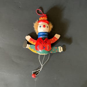 May include: A vintage wooden clown doll with a red jacket, blue collar, and a hat that says "LILLIA". The clown has outstretched arms and legs, with the year "1994" on one foot. A red string is attached to the clown.