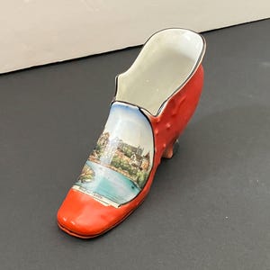 May include: A ceramic shoe-shaped vase in bright red with a detailed illustration of Windsor Castle. The shoe has a white interior and a small heel. The illustration is on the front of the shoe.