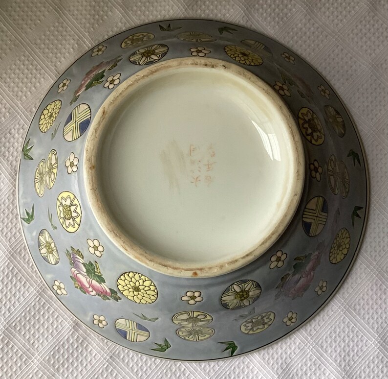 May include: A large, blue and white porcelain bowl with a floral and geometric design. The bowl has a round shape and a flat bottom. The bottom of the bowl has a mark in Japanese characters.