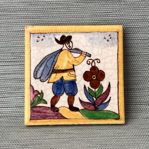 May include: A hand-painted ceramic tile featuring a whimsical illustration of a person in a yellow shirt and blue pants carrying a blue bag and playing a musical instrument. The tile has a yellow border and a white background with a brown flower.