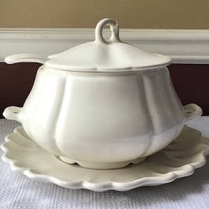 May include: White ceramic soup tureen with a lid and a matching serving plate. The tureen has a rounded, bulbous shape with a fluted design.
