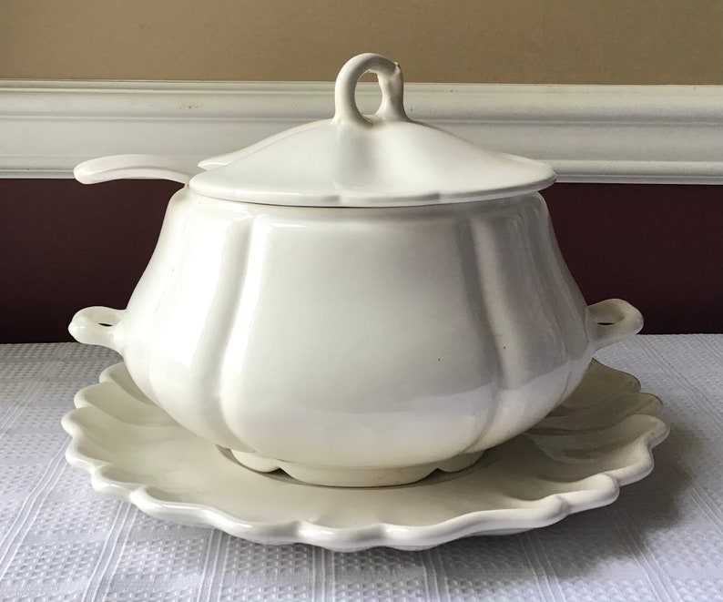 May include: White ceramic soup tureen with a lid and a matching serving plate. The tureen has a rounded, bulbous shape with a fluted design.