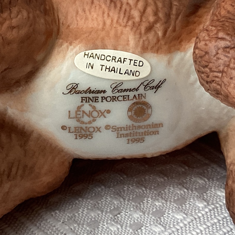 May include: A fine porcelain figurine of a Bactrian camel calf. The figurine is tan and white, with a Lenox and Smithsonian Institution stamp. The text on the figurine reads "Handcrafted in Thailand" and "Fine Porcelain."