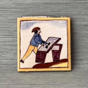 May include: A hand-painted ceramic tile featuring a cartoon illustration of a person in blue clothing pushing a brown and purple box. The tile has a yellow border and a white background.