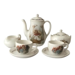May include: A white ceramic tea set featuring a teapot, sugar bowl, creamer, and two teacups with saucers. The set is decorated with a whimsical illustration in muted tones. The teapot has a lid and a curved spout.