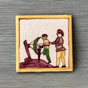 May include: A hand-painted ceramic tile depicting two figures operating a hand-cranked grinding wheel. The tile has a yellow border and a white background with a purple and green landscape.