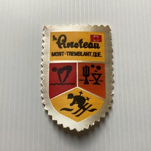 May include: A vintage fabric patch with a shield design. The patch features the text "Le Pinoteau MONT-TREMBLANT, QUE." and various icons representing activities. The patch has a gold border and a cream-colored background.