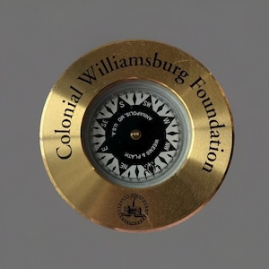 May include: A brass compass with the text "Colonial Williamsburg Foundation" around the edge. The compass face is black and white, with directional markings and the text "Annapolis, MD, U.S.A."