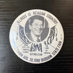 May include: A round, white button with a black and white image of Ronald Reagan. Text includes "Illinois is Reagan Country," "Republican," and "Duquion State Fair." The button also lists his birth date and place.