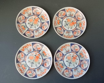 May include: Set of four white ceramic plates with a floral design. The plates feature a central motif of a tree with orange flowers and green leaves, surrounded by a repeating pattern of blue and orange floral elements. The plates have a white background with a gold rim.