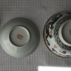 May include: A set of three white ceramic bowls with a dragon design. The bowls have a red and green design with a gold trim. The bottom of the bowls have the text "Made in China" in red.