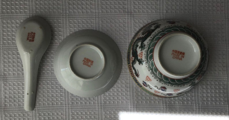 May include: A set of three white ceramic bowls with a dragon design. The bowls have a red and green design with a gold trim. The bottom of the bowls have the text "Made in China" in red.