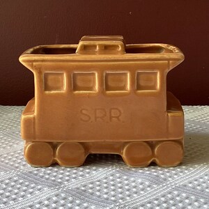 May include: A brown ceramic train engine planter with the letters "SRR" on the side. The planter has a small roof and four wheels.